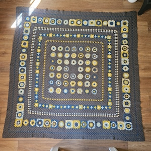May include: A square crochet blanket featuring a dark gray base with a central grid of small floral motifs in shades of yellow, light blue, and white. The blanket is framed by concentric borders of dark gray yarn and decorative square motifs.
