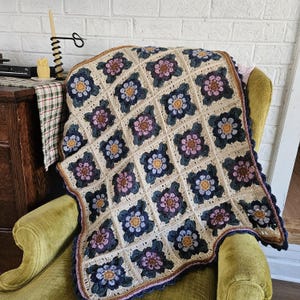 May include: A handmade crochet blanket with a floral pattern. The blanket features square blocks with blue, pink, and yellow flowers on a cream background. The blanket is draped over a green armchair, showcasing its intricate design and craftsmanship.
