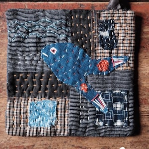 May include: A handmade textile square with a blue fish applique. The square is composed of various fabric patches in shades of blue, black, and brown, with visible stitching. A leather loop is attached to the top.