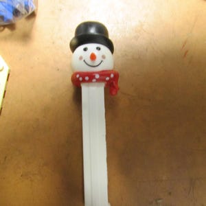 May include: A white plastic snowman-shaped candy dispenser. The snowman has a black top hat, a red scarf with white polka dots, and a smiling face with a carrot nose. The dispenser is a novelty item.