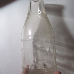May include: A clear glass bottle with a narrow neck and a wide base. The bottle has a cylindrical shape with a slightly rounded shoulder. The word "SMITH" is embossed on the front of the bottle. The bottle is held by a hand.