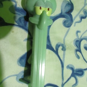 May include: A light green Squidward Tentacles PEZ dispenser. The dispenser features Squidward's face with yellow eyes and a long nose. The dispenser is made of plastic and has a long, rectangular body.