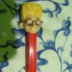 May include: A vintage Pez dispenser featuring a cartoon character with yellow hair, round glasses, and a smiling face. The dispenser has a red body and base. The background has a blue and white floral pattern.