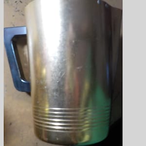 May include: A gold-colored metal pitcher with a black handle. The pitcher has horizontal ridges near the bottom. The handle is rectangular and attached to the side of the pitcher. The pitcher is empty.
