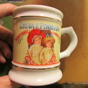 May include: A vintage ceramic mug with a cream-colored background and gold trim. The mug features an illustration of two children in hats and the text "LYDIA E. PINKHAM VEGETABLE COMPOUND". The handle is white.