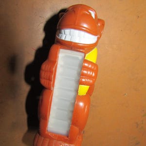 May include: A vintage orange plastic toy shaped like a smiling cartoon character with a large white-toothed grin. The toy has a rectangular opening with a series of white plastic steps. A yellow accent is visible on the side.