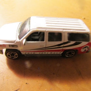 May include: A white toy SUV with black and red graphics on the side. The toy car has black wheels with a star design and a detailed front end. The vehicle has a red and black design on the rear.