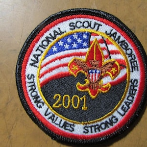 May include: A circular embroidered patch featuring the text National Scout Jamboree and Strong Values Strong Leaders with the year 2001 in yellow. The design includes a red, white, and blue American flag background and a central gold fleur-de-lis.