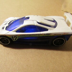 May include: A white and blue toy car with black wheels. The car has a spoiler and the text "PROTOTYPE HW" and the number "12" on the side. The car is a miniature replica of a sports car.