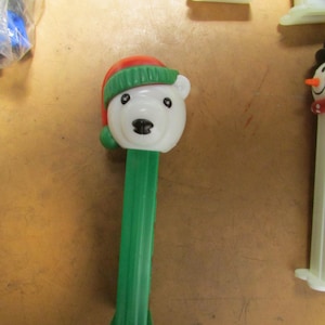 May include: A white polar bear Pez dispenser wearing a red and green hat. The dispenser has a green stem and is part of a collection of novelty candy dispensers. The bear's face has black eyes and a black nose.