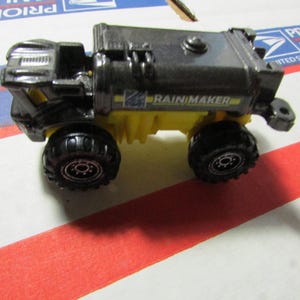 May include: A black and yellow toy truck with large black tires. The truck has the words "RAINMAKER" printed on the side. The toy truck is on a white surface with red and blue stripes.