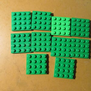 May include: A collection of eight square green plastic building plates, each featuring a four-by-four grid of circular studs on the top surface. These modular construction components are arranged in a loose cluster, displaying a uniform bright green color and a consistent geometric design across all individual pieces.