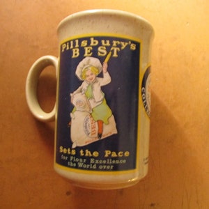May include: Vintage Pillsbury's Best coffee mug. The mug features a blue and white graphic of the Pillsbury Doughboy with the text "Pillsbury's Best" and "Sets the Pace for Flour Excellence the World over". The mug has a beige handle and rim.