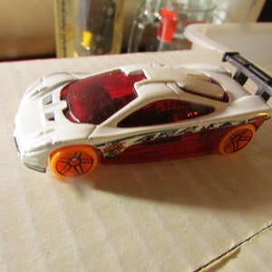 May include: A white and red toy race car with orange wheels and a black spoiler. The car has a red tinted window and a detailed design on the side. This toy car is a collectible item.