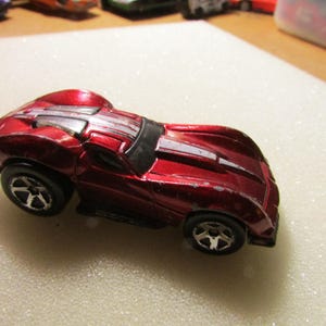 May include: A red toy car with silver stripes on the hood and roof. The car has black wheels with silver rims. The toy car is a vintage collectible.