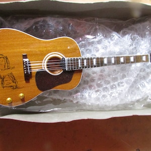 May include: A miniature acoustic guitar with a light brown finish, featuring a white soundhole ring and a dark brown pickguard. The guitar has a black headstock and a fretboard with white inlays. The body is adorned with sketches of four faces and signatures.