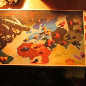 May include: Vintage Risk board game featuring a world map with continents and oceans. The game board shows territories with red and orange game pieces. A cannon graphic is on the left side of the board, and the word "RISK" is in large red letters on the right.