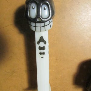 May include: A black and white skull-shaped Pez dispenser. The skull has large, oval eyes and a wide grin. The dispenser's stem is white with a black bone design. A fun and collectible novelty item.