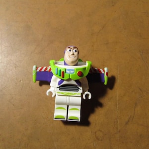 May include: A Buzz Lightyear minifigure featuring a white and green space suit with purple wings, a printed chest control panel, and a molded head with a smiling expression. The character has articulated arms and legs with green accents.