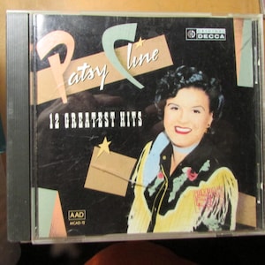 May include: A Patsy Cline 12 Greatest Hits music CD in a clear plastic jewel case. The cover features a portrait of the artist wearing a black shirt with yellow fringe and floral accents, set against a black background with geometric shapes and stars.