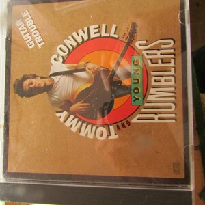 May include: A CD case featuring the album "Guitar Trouble" by Conwell and Tommy Young Rumblers. The cover art shows a man playing a guitar, set against a brown background with a red and orange circular design. The text is in white and black.