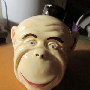 May include: A ceramic monkey head figurine with a black top hat. The monkey has a cream-colored face, blue eyes, and a red smiling mouth. The figurine is a vintage collectible.