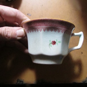 May include: A small, antique teacup with a white base and a burgundy and yellow rim. The cup features a floral design and a decorative burgundy pattern around the rim. The cup is held by a hand.