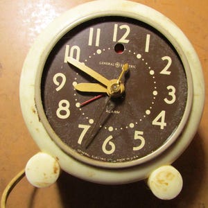 May include: Vintage General Electric alarm clock with a cream-colored casing and a brown face. The clock features gold-colored hands and white numerals. The text "GENERAL ELECTRIC" and "ALARM" are visible on the face. Made in the USA.