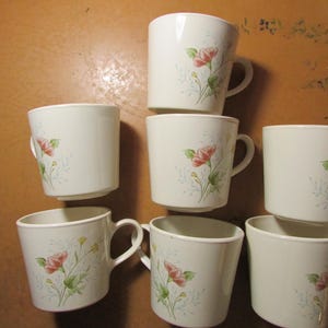 May include: A collection of seven white ceramic mugs. Each mug features a floral design with pink flowers and green leaves. The mugs have a simple, rounded shape with a handle on the side. The mugs are arranged on a brown surface.