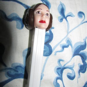 May include: A white PEZ dispenser featuring a character head with brown hair and red lips. The character's face is detailed with painted eyes and eyebrows. The dispenser is set against a blue and white patterned background.