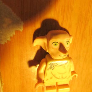 May include: A tan plastic toy figure with large pointed ears, a long nose, and green eyes. The figure has a printed design on the torso resembling a simple garment with a tie at the neckline. It features standard articulated arms and legs.