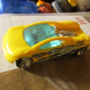May include: Yellow toy car with blue tinted windows and flame decals. The car has a unique design with a visible engine and a spoiler. The wheels are black and silver. The car is sitting on a white surface.