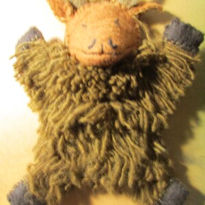 May include: A handmade plush toy shaped like a highland cow with a tan felt face, two small horns, and a shaggy olive green yarn body. The toy features dark gray felt hooves at the ends of its four limbs and simple stitched facial features.