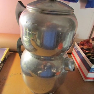 May include: A vintage, stainless steel coffee percolator with a black handle and lid knob. The percolator has a two-part design with a spout and a lid. The exterior is shiny and reflective, showing the surrounding environment.