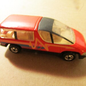 May include: A red toy van with a black roof and clear windows. The van has a yellow and pink lightning bolt design on the side, with blue and orange stripes. The wheels are black.