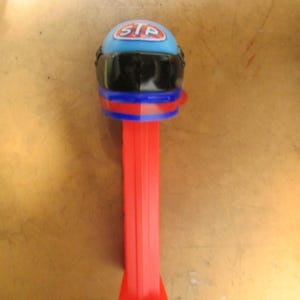 May include: A red, blue, and black PEZ dispenser shaped like a racing helmet. The helmet is blue with the letters "STP" in white. The dispenser has a red stem.