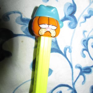 May include: A Garfield PEZ dispenser featuring the cartoon cat wearing a blue nightcap. The dispenser has an orange head, a yellow stem, and a grumpy expression. The background is a white and blue floral pattern.