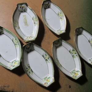 May include: A set of six white porcelain dishes with an elongated, boat-like shape. Each dish features a delicate floral pattern with yellow and green accents, finished with a thin gold trim along the edges and a green border detail.