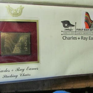 May include: A framed stamp and envelope commemorating Charles and Ray Eames. The stamp features a gold design, while the envelope includes a chair illustration and the text "First Day of Issue." The text "Charles + Ray Eames Stacking Chairs" is also visible.