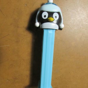 May include: A blue and black penguin PEZ dispenser. The penguin has a light blue hat with white pom-poms and a white ball on top. The dispenser's stem is light blue.