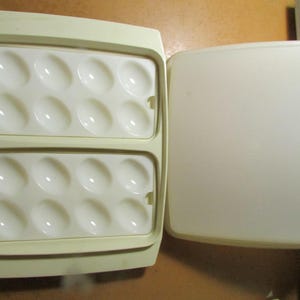 May include: A cream-colored plastic egg storage container featuring two removable white trays with eight oval-shaped indentations each. The set includes a matching cream-colored lid that covers the base, providing a secure enclosure for eggs.