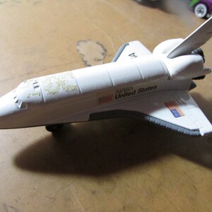 May include: A white toy space shuttle with gray accents, featuring the NASA and United States flag logos. The toy has a detailed design with wings and a tail fin. The shuttle is on a brown surface.