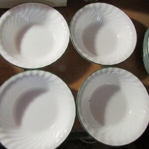 May include: Four white ceramic bowls with a green trim and a spiral design. The bowls are arranged in a square formation, with additional plates visible in the background. The plates also have a green trim.