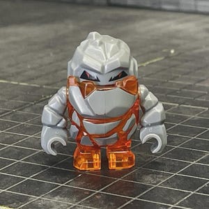 May include: A small, stylized toy figure constructed from gray, rock-like segments with a jagged, textured head and torso. The figure features translucent orange accents integrated into the chest and legs, with sharp, dark red eyes set into the gray face.