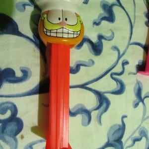 May include: A vintage orange and white Garfield PEZ dispenser. The dispenser features Garfield's face with a chef's hat and a large, yellow mustache. The dispenser's stem is red.