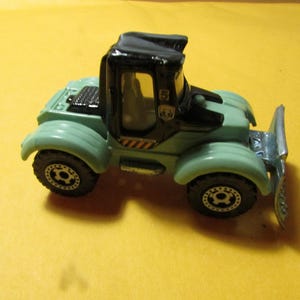 May include: A die-cast toy tractor in light green and black with a silver front plow attachment. The vehicle features a black cab with the number 5 printed on the side, black wheels with silver rims, and yellow and black hazard stripes on the side.