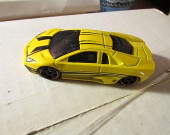 May include: A yellow toy car with black accents and black wheels. The car has a sleek design with a dark tinted windshield and windows. The toy car is a miniature replica of a sports car.