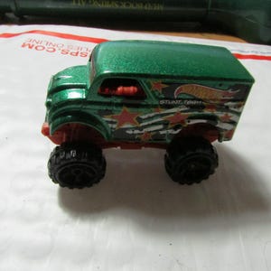 May include: A green and red Hot Wheels Stunt Team toy truck with black wheels. The truck has a green roof and is decorated with stars and stripes. The text "STUNT TEAM" is visible on the side.