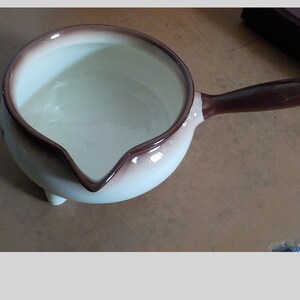 May include: A small, round, white and brown ceramic saucepan with a long handle. The interior is white, and the rim and handle are brown. The saucepan has a spout and three small feet.