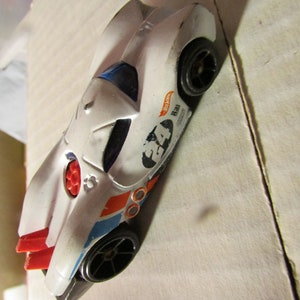May include: White toy race car with black wheels, red and blue accents, and the number 34. The car has a sleek design with a blue windshield and the Hot Wheels logo.